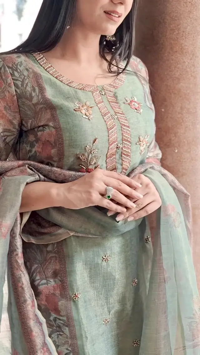Close view of a sage-green kurta with a pink floral print and hand embroidery at the neckline, a matching dupatta held in one hand.